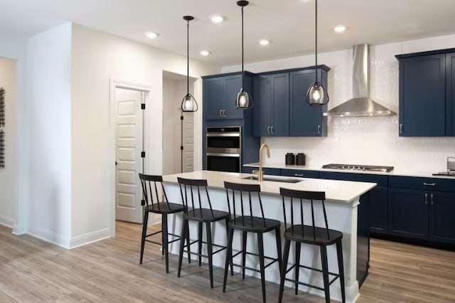 kitchen featuring wall chimney exhaust hood, an island with sink, sink, decorative light fixtures, and blue cabinets