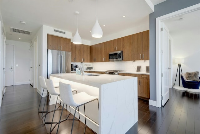 kitchen with appliances with stainless steel finishes, dark hardwood / wood-style flooring, hanging light fixtures, and an island with sink