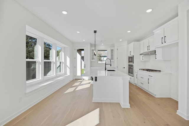 kitchen with stainless steel appliances, pendant lighting, white cabinets, light hardwood / wood-style floors, and an island with sink