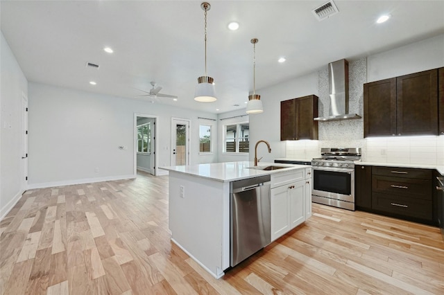 kitchen with light hardwood / wood-style floors, appliances with stainless steel finishes, a center island with sink, and wall chimney range hood