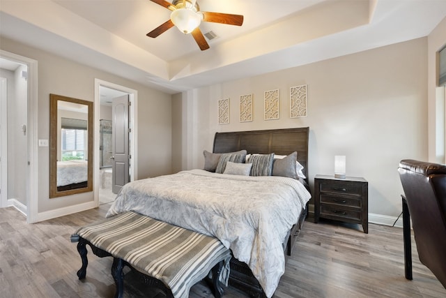 bedroom featuring light hardwood / wood-style floors, ceiling fan, ensuite bath, and a tray ceiling