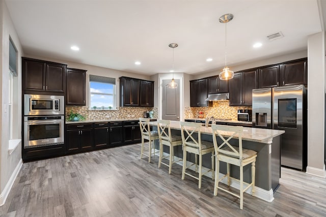 kitchen with appliances with stainless steel finishes, a kitchen island with sink, light hardwood / wood-style floors, and hanging light fixtures