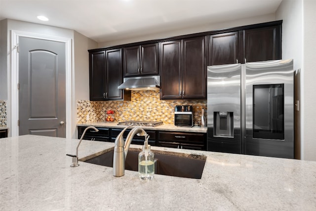 kitchen with sink, tasteful backsplash, light stone countertops, dark brown cabinets, and appliances with stainless steel finishes