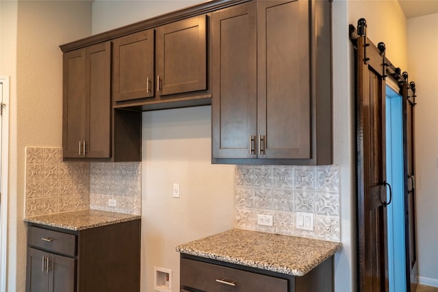 kitchen featuring backsplash, a barn door, dark brown cabinets, and light stone countertops