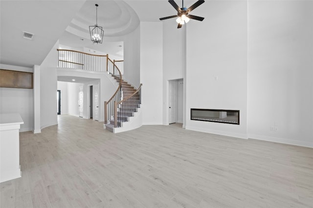 unfurnished living room featuring a tray ceiling, a towering ceiling, light hardwood / wood-style floors, and ceiling fan with notable chandelier