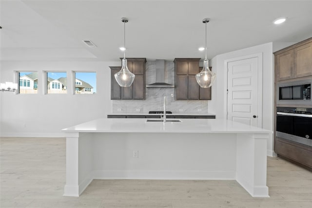 kitchen featuring built in microwave, stainless steel oven, wall chimney exhaust hood, backsplash, and an island with sink