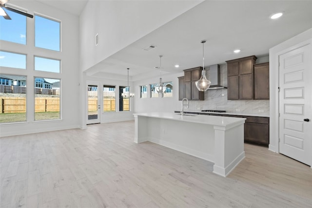 kitchen with tasteful backsplash, wall chimney range hood, sink, hanging light fixtures, and an island with sink