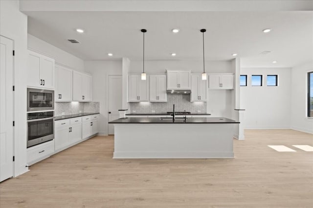 kitchen with oven, white cabinetry, hanging light fixtures, and an island with sink