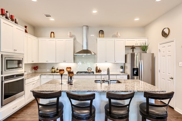 kitchen with a center island with sink, sink, white cabinetry, appliances with stainless steel finishes, and wall chimney range hood