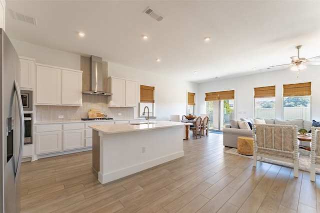 kitchen with light wood-type flooring, a kitchen island with sink, wall chimney exhaust hood, and white cabinets