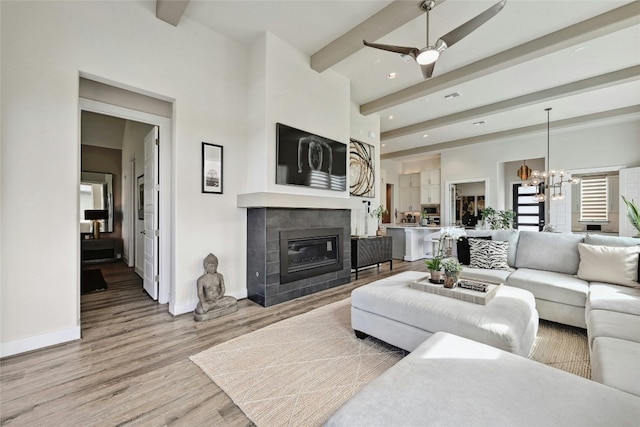 living room featuring hardwood / wood-style floors, a tiled fireplace, beamed ceiling, and ceiling fan with notable chandelier