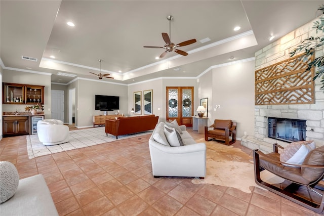 living room with a stone fireplace, ceiling fan, beverage cooler, and a tray ceiling