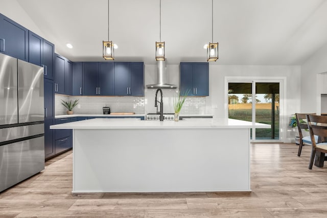 kitchen featuring blue cabinets, a kitchen island with sink, stainless steel refrigerator, pendant lighting, and light wood-type flooring