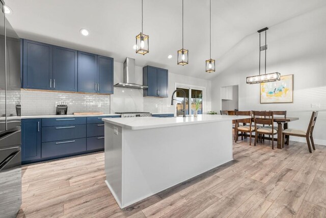 kitchen with an island with sink, wall chimney range hood, pendant lighting, and blue cabinets