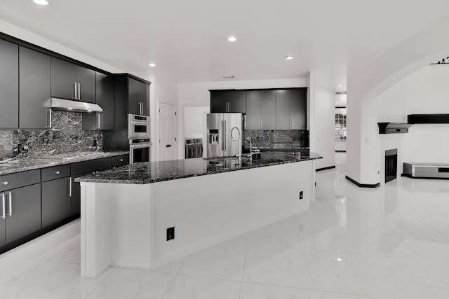 kitchen featuring stainless steel appliances, dark stone countertops, a kitchen island with sink, and tasteful backsplash