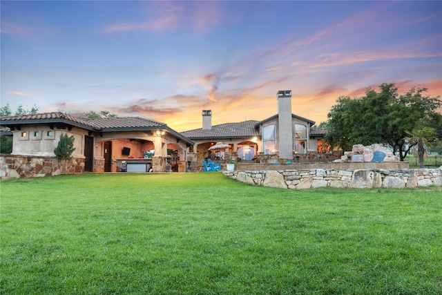 back of property at dusk with a tile roof, stone siding, a lawn, stucco siding, and a chimney