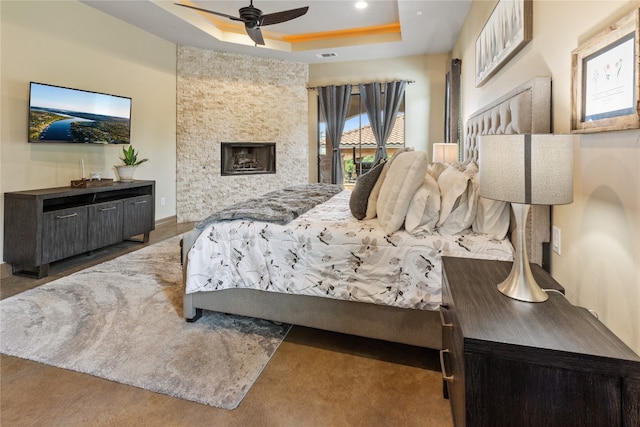 bedroom with a stone fireplace, dark wood-type flooring, ceiling fan, and a raised ceiling