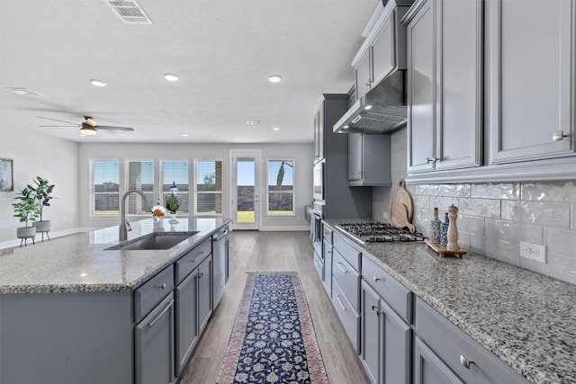 kitchen with gray cabinetry, sink, an island with sink, appliances with stainless steel finishes, and light hardwood / wood-style floors