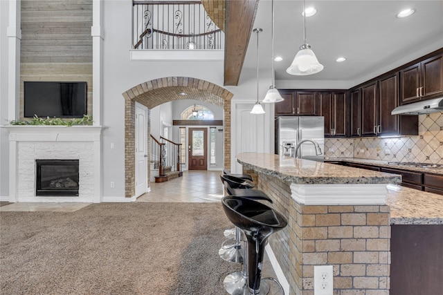 kitchen with a center island with sink, stainless steel appliances, dark brown cabinets, light stone countertops, and pendant lighting