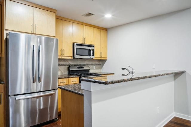 kitchen with dark wood-type flooring, stainless steel appliances, kitchen peninsula, dark stone counters, and decorative backsplash