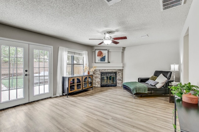 living area with a stone fireplace, french doors, a textured ceiling, ceiling fan, and light wood-type flooring