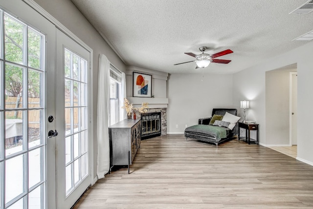 living area featuring a stone fireplace, french doors, a textured ceiling, ceiling fan, and light wood-type flooring