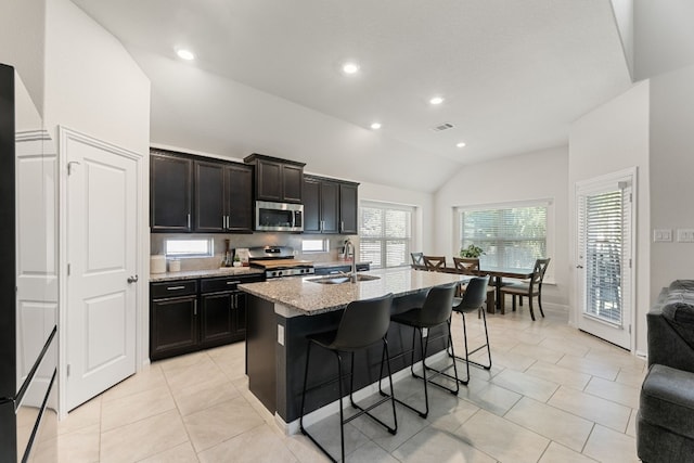 kitchen featuring stainless steel appliances, a center island with sink, vaulted ceiling, sink, and light stone countertops