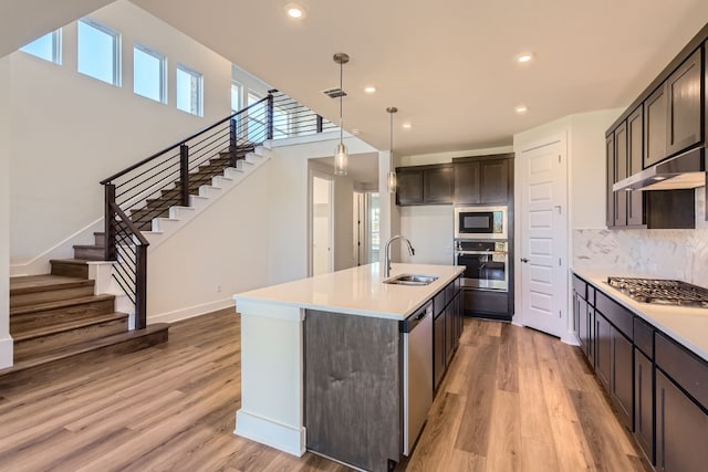 kitchen featuring stainless steel appliances, an island with sink, pendant lighting, decorative backsplash, and light wood-type flooring