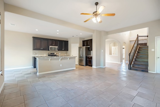 kitchen featuring arched walkways, baseboards, an island with sink, ceiling fan, and stainless steel appliances