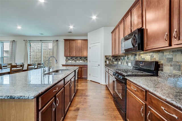 kitchen with black appliances, light wood-type flooring, light stone countertops, sink, and a kitchen island with sink