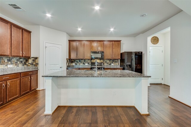 kitchen with a center island with sink, black appliances, dark wood-type flooring, and backsplash