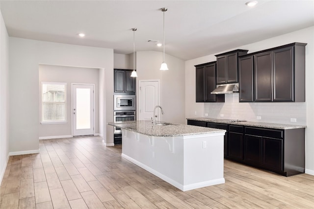 kitchen featuring vaulted ceiling, hanging light fixtures, a kitchen island with sink, light stone countertops, and appliances with stainless steel finishes