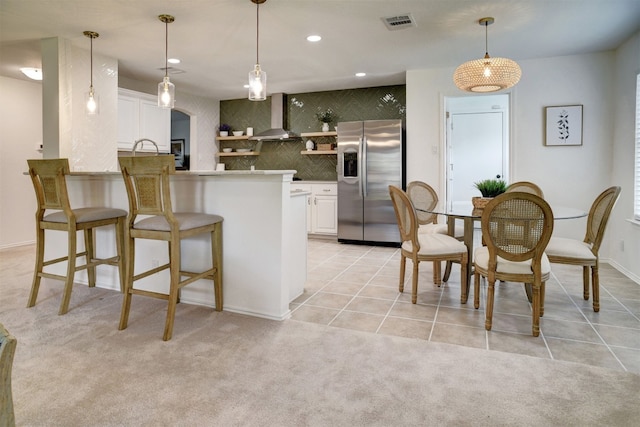 kitchen with kitchen peninsula, light carpet, wall chimney range hood, white cabinetry, and stainless steel fridge