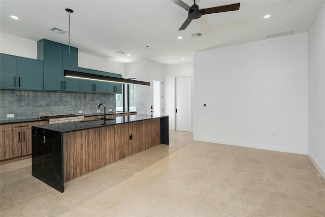 kitchen with a center island with sink, backsplash, decorative light fixtures, sink, and dark stone countertops