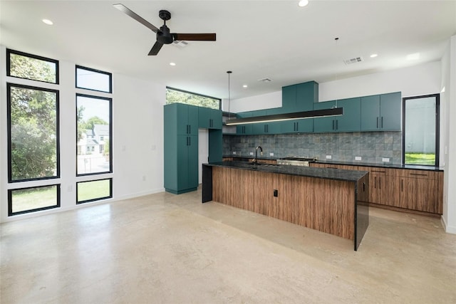 kitchen with sink, a kitchen island with sink, backsplash, pendant lighting, and dark stone countertops