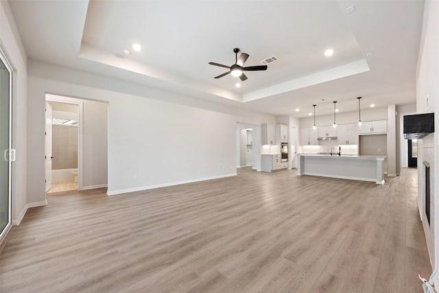 unfurnished living room with a raised ceiling, ceiling fan, sink, and light hardwood / wood-style floors