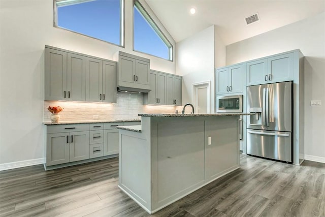 kitchen featuring built in microwave, light stone countertops, high vaulted ceiling, stainless steel fridge, and an island with sink