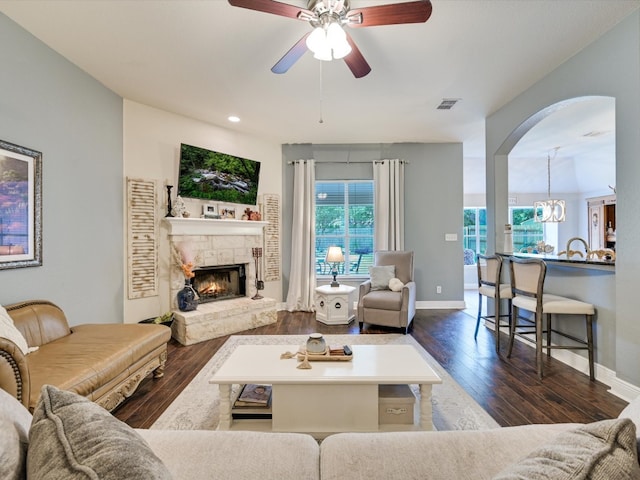 living room featuring dark wood-type flooring, a stone fireplace, and ceiling fan with notable chandelier