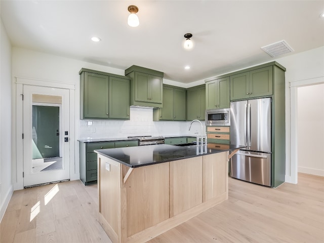 kitchen with green cabinetry, decorative backsplash, an island with sink, light wood-type flooring, and appliances with stainless steel finishes