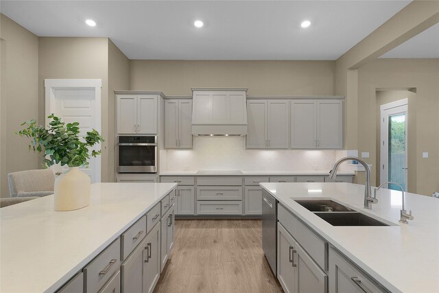 kitchen with appliances with stainless steel finishes, sink, premium range hood, light wood-type flooring, and tasteful backsplash