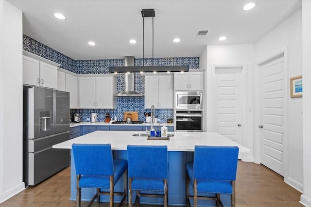 kitchen featuring white cabinetry, wall chimney range hood, stainless steel appliances, an island with sink, and hanging light fixtures