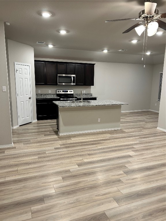 kitchen featuring stainless steel appliances, light hardwood / wood-style floors, light stone countertops, and an island with sink