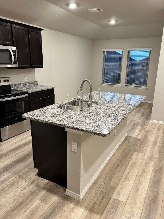 kitchen with light stone counters, stainless steel appliances, light wood-type flooring, sink, and a kitchen island with sink