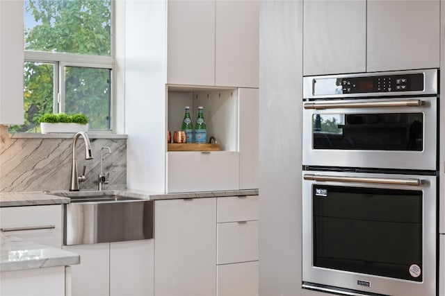 kitchen with stainless steel double oven, white cabinetry, light stone countertops, sink, and decorative backsplash