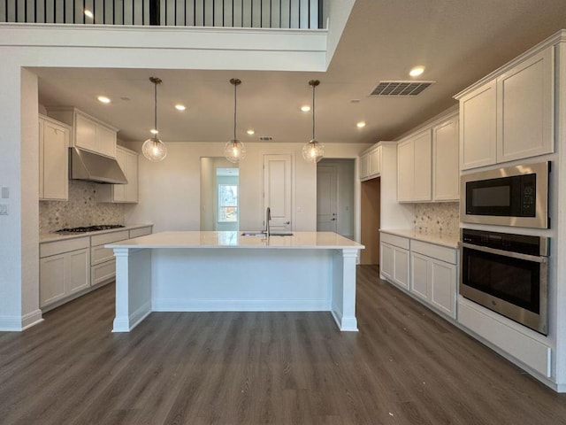 kitchen with under cabinet range hood, built in microwave, a center island with sink, stainless steel oven, and dark wood-style floors