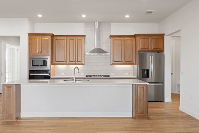 kitchen featuring wall chimney exhaust hood, light hardwood / wood-style flooring, sink, a kitchen island with sink, and appliances with stainless steel finishes