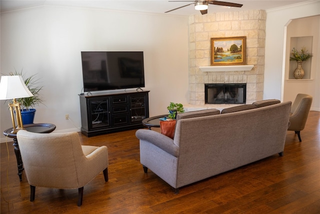 living room featuring a stone fireplace, dark wood-type flooring, ceiling fan, and crown molding