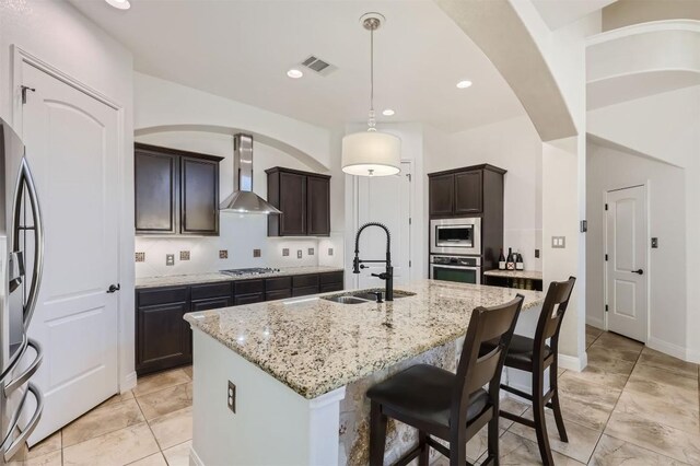 kitchen with stainless steel appliances, dark brown cabinetry, an island with sink, wall chimney range hood, and decorative light fixtures