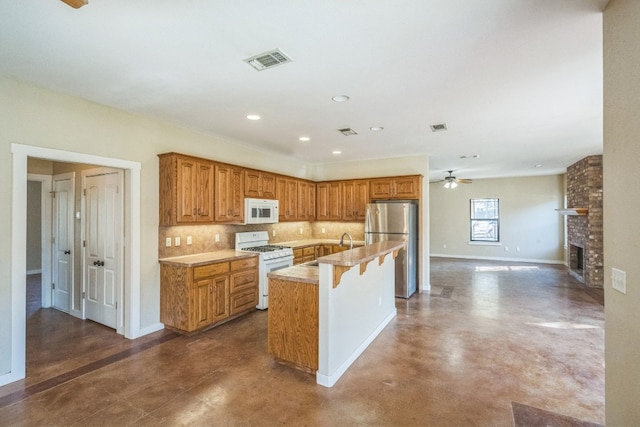 kitchen with a brick fireplace, a center island with sink, tasteful backsplash, a kitchen breakfast bar, and white appliances