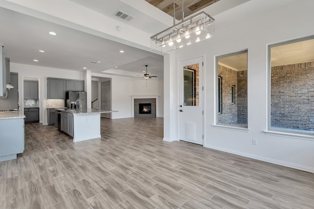 kitchen with stainless steel refrigerator with ice dispenser, light wood-type flooring, gray cabinetry, a kitchen island with sink, and pendant lighting